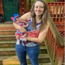 Whitney smiles in a red, white, and blue blouse with blue jeans. She is holding a baby.
