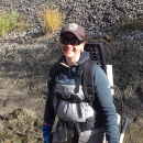 A smiling woman in sunglasses, Service ballcap, fleece jackets, and waders wears a backpackable electroshocker box on her back and holds two rods in her hands for carrying the current to the water.