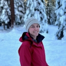 woman in white hat and red jacket in front of a snowy wooded background.
