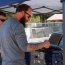 A man with sleeves rolled up interacts with a computer outside under a canopy.