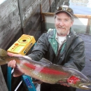 A man in rainjacket and ballcap stands in a concrete tank, holding up in both hands a gorgeous steelhead in bright spawning colors.