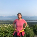 Woman in pink shirt stands at overlook with vegetation behind her and view of bay and mountains.