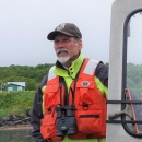 man in usfws hat and lifejacket steers a skiff with hill and houses on the shore in the background