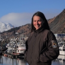 woman in a USFWS uniform stands in front of a boat harbor with mountains in the background