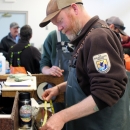 A man in Service jacket, waders, and ballcap measures a fish in a busy room with other similarly dressed people.