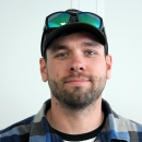 A lightly bearded man with ballcap, sunglasses perched on the camp, and a plaid flannel shirt stands in front of a white wall for a photo.