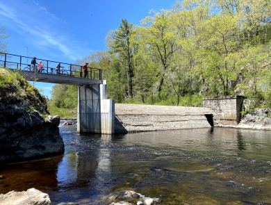 A dam crosses a river, but a portion has been removed to that water can flow through. People stand on an overlook that crosses the open section of the river. 