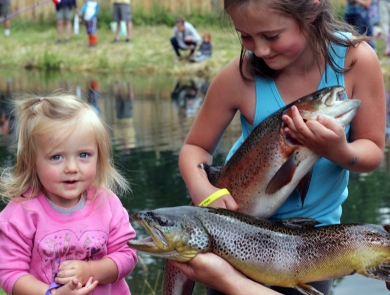 Two girls pose with two big trout in front of a pond where other children are fishing.