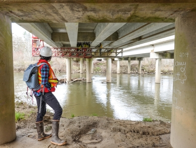 Person standing on a river bank beneath a bridge watching a crew working on a catwalk