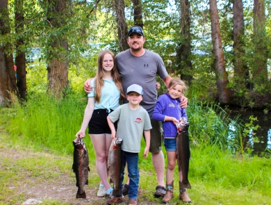 A man with two girls and a boy stand in front of trees, all 3 kids holding huge trout.