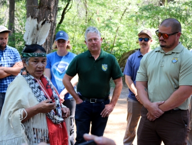 A woman in traditional Native American clothing speaks with several people wearing federal- or state-agency shirts and hats