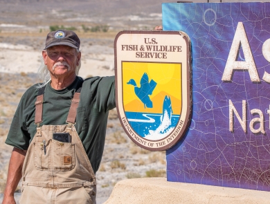 A man with a white mustache wearing overalls and a U.S. Fish and Wildlife Service volunteer cap standing next to a sign with the U.S. Fish and Wildlife Service logo on it