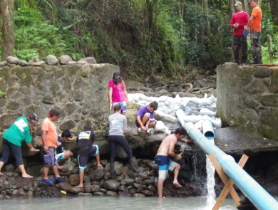 Several students in various colored clothes assist with fishway construction. A large tube stems up from a river to a smaller stream that is being directed with stones.