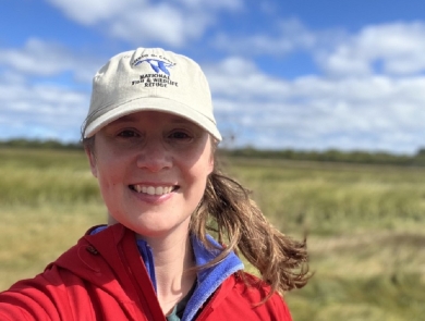 Woman stands in a marsh wearing fish and wildlife hat