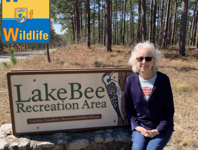 Deb Adams sits on the welcome sign at Lake Bee Recreation area at Carolina Sandhills National Wildlife Refuge. The sign has a woodpecker carved into it. Deb is wearing sunglasses and smiling. The WoW: Woman of Wildlife logo is in the upper left corner. 