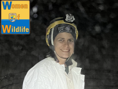 Pam McDill smiles at the camera as she stands in dark cave. She is wearing a white jumpsuit and a helmet with a headlamp on it. The WoW: Women of Wildlife logo is in the upper left corner of the image.