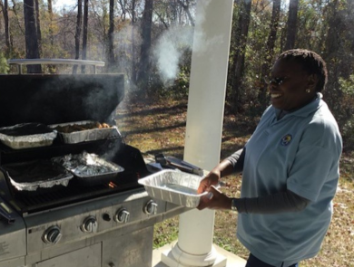 Brenda Williams, volunteer at Waccamaw National Wildlife Refuge, smiles as she holds a tray and stands near a grill where food is being cooked.