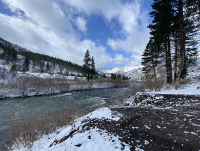 An image of a river flowing thorugh a mountain canyon dusted with snow and pine trees on either side under a partly cloudy blue sky. 