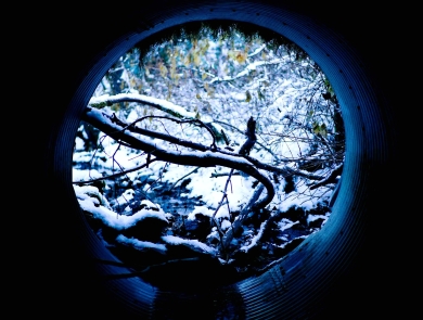 looking out at a snowy stream from within a round metal culvert pipe.