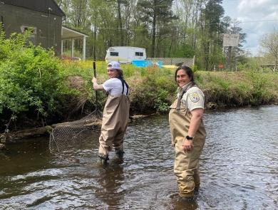 Two people standing in a river wearing waders. One is holding a large net to capture fish in. 