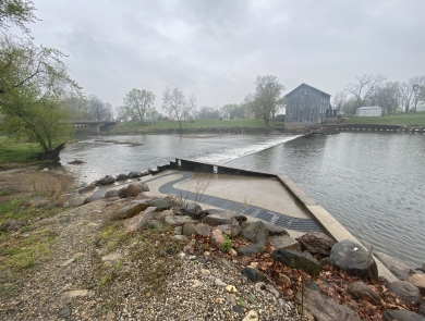 A metal fish ladder on a concrete slope next to a low-head dam near the Stockdale Mill on a cloudy day.