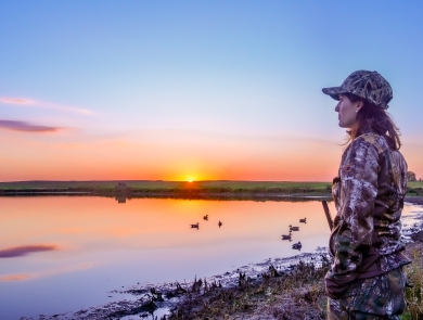 A woman waterfowl hunting by a wetland at sunset