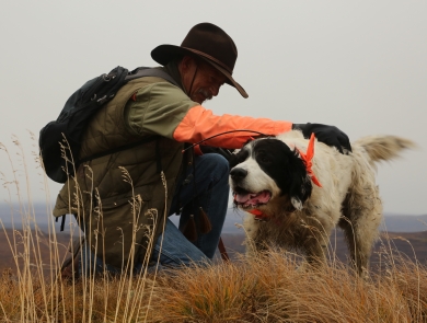 A hunter wearing blaze orange sleeved shirt and hat kneels down touching a hunting dog on a grassy hill top with out of focus hills in the background. 