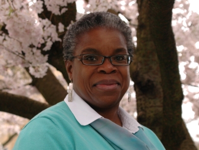A woman wearing glasses and a blue sweater smiles while standing under a blooming cherry blossom tree