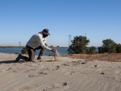 Kneeling man picks up a handful of sand with a body of water in the background