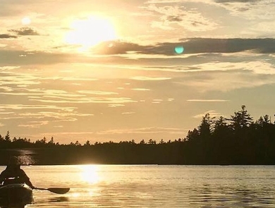 A kayaker paddling toward a setting sunset on a pond surrounded by evergreen trees