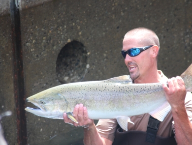 A man in waders and sunglasses holds up a large salmon horizontally with both hands.