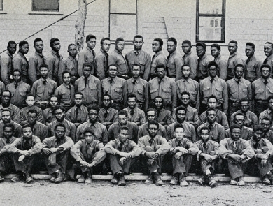 Black and white photo of more than 100 young African American men in uniform posing in front of a building