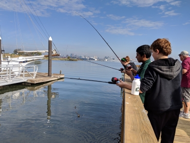 Three students fish from pier at Masonville Cove with Baltimore skyline in background