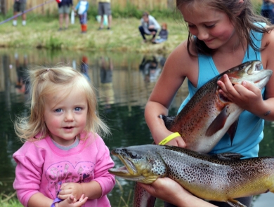 A very young blond-haired girl and an older girl pose with large trout in front of a pond where other children are fishing.