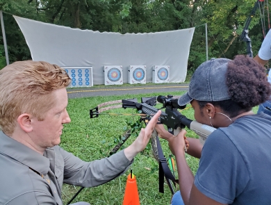 A man teaching a woman how to handle a crossbow while she aims at an archery target