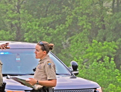 Two law enforcement officers -- a man and a woman -- speaking as the male officer points. They are in front of a police car in a wooded setting.
