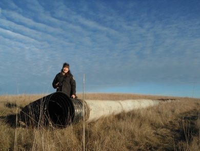 An uniformed adult stands in front of one end of a long drain pipe in a grassy prairie