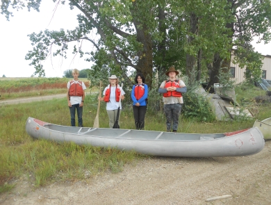 4 youth standing in a line behind an aluminum canoe outside