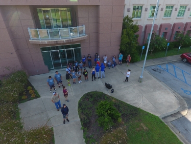 An aerial photograph of a group of people standing on the sidewalk outside of a large building