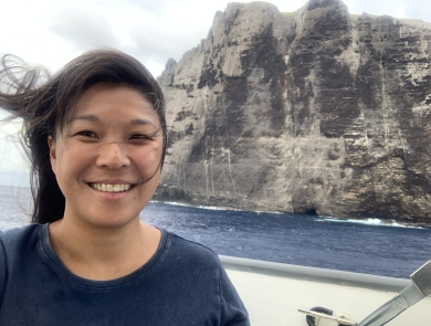 Jiny Kim, wearing a navy blue shirt, stands on a boat with Nihoa in the background.