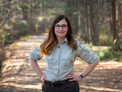 A woman with long brown hair stands on a trail framed by forest. Her hands are on her hips. She wears glasses and a U.S. Fish and Wildlife Service Uniform. 