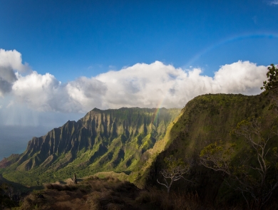 A view of mountains on Kauai where the mitigation site is. The clouds break behind the mountain cliffs and a rainbow arches in front.