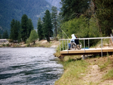 A man wearing a hoodie and sitting in a wheelchair fishes from a platform overhanging a river.