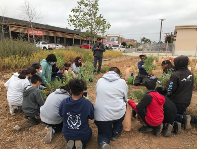 A group of students kneel next to each other in a semi circle as their teacher stands on the left end of the semi circle. Each student has a single potted plant in front of them. They are either looking at their plant or listening to their instructor who is also in a kneeling-sitting position holding out a green and shrubby potted plant. 