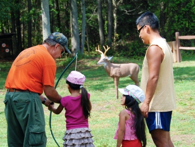 man helping girl learn archery. Another man and girl look on.