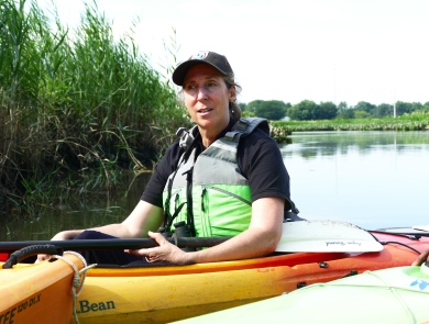 woman in lifevest in kayak in water near a shore with tall grasses