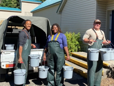Two women and one man stand smiling in the sun in waders holding blue buckets.