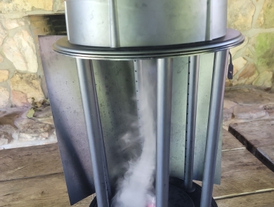 Two foot high hollow metal cylinder 10 inches in diameter sitting on top of a wood picnic table. There is tan mortared stonework behind the table and a youth hand next to the machine. The hollow cylinder is open in the front and smoke is swirling in an upside down vortex. 