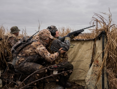 Person is camouflage hunting gear crouches behind a second person in a wheelchair in a duck hunting blind