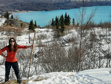 A woman standing wearing a bright orange shirt and sunglasses holding a walking stick in a snowy landscape. A bright, aqua blue lake in the background.